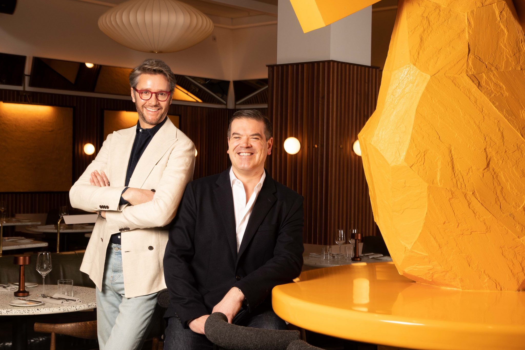 Two men posing in a trendy restaurant with modern decor and a large yellow geometric sculpture in the background.
