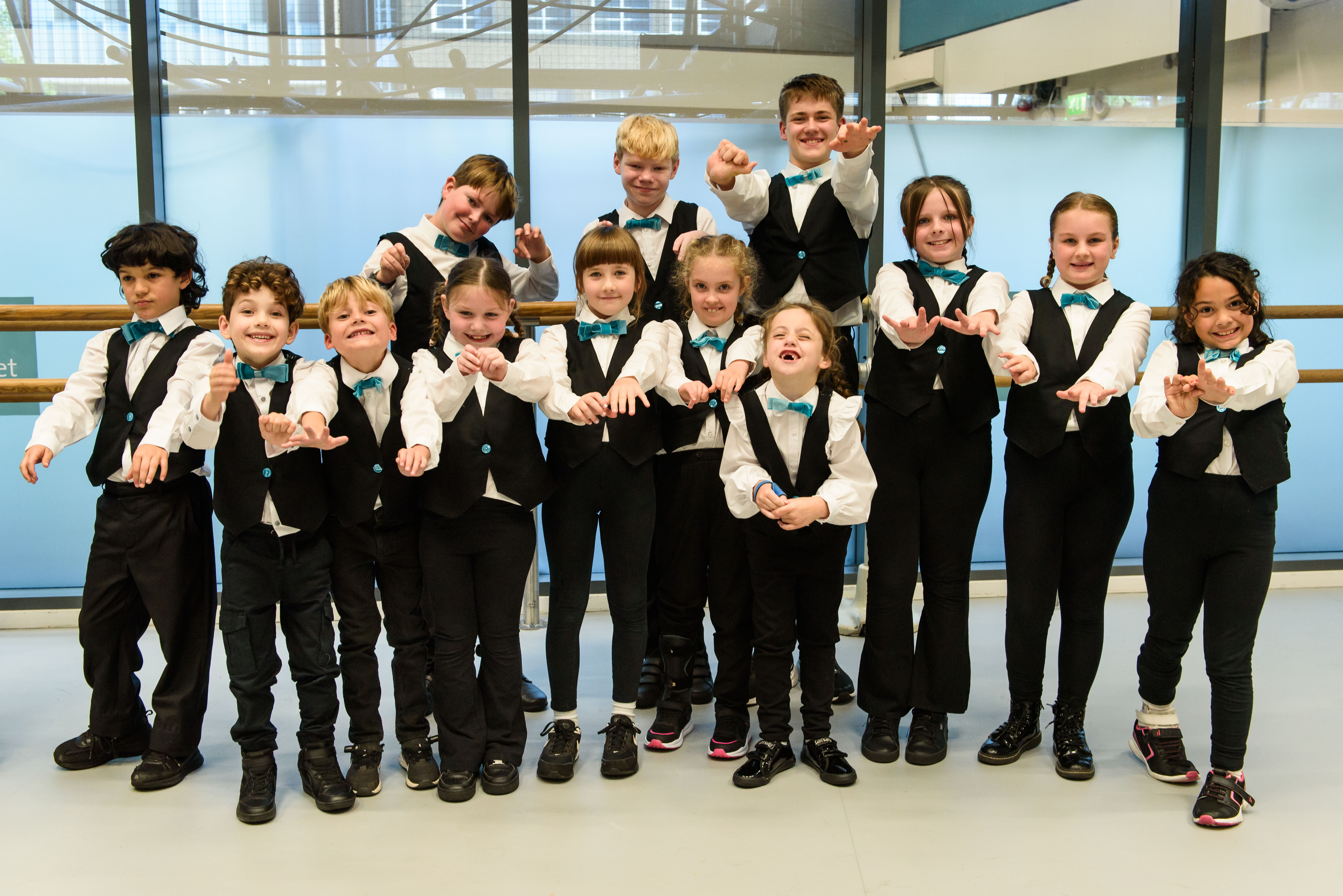 Group of kids in matching vests and teal bow ties posing with playful hand gestures.