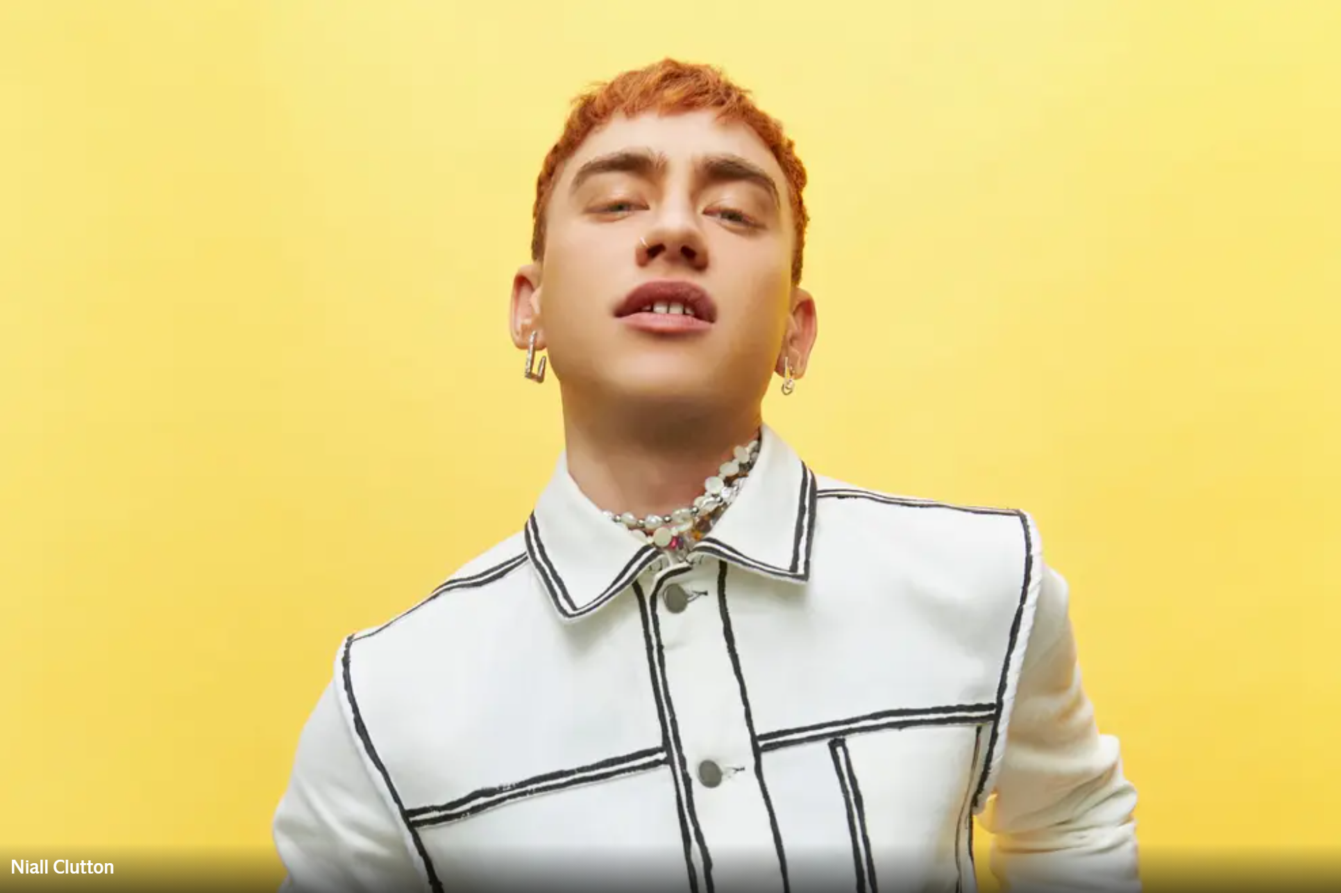 Portrait of olly alexander with orange hair, wearing a stylish white jacket, set against a bright yellow background