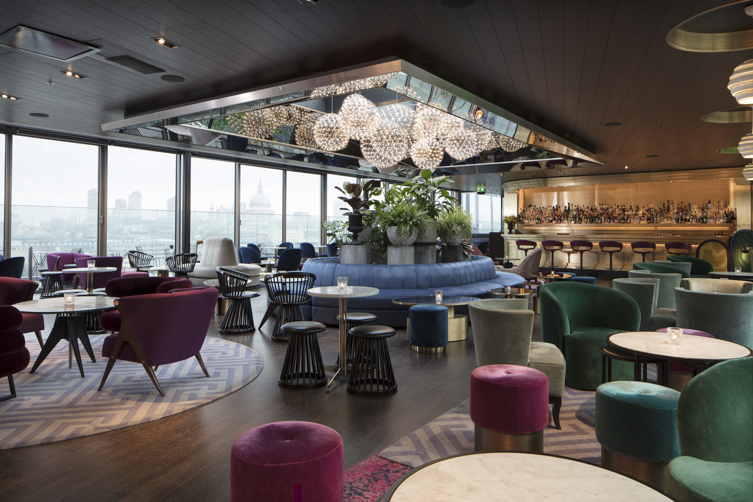 A lively rooftop bar with vibrant seating, lush greenery, and panoramic city views.