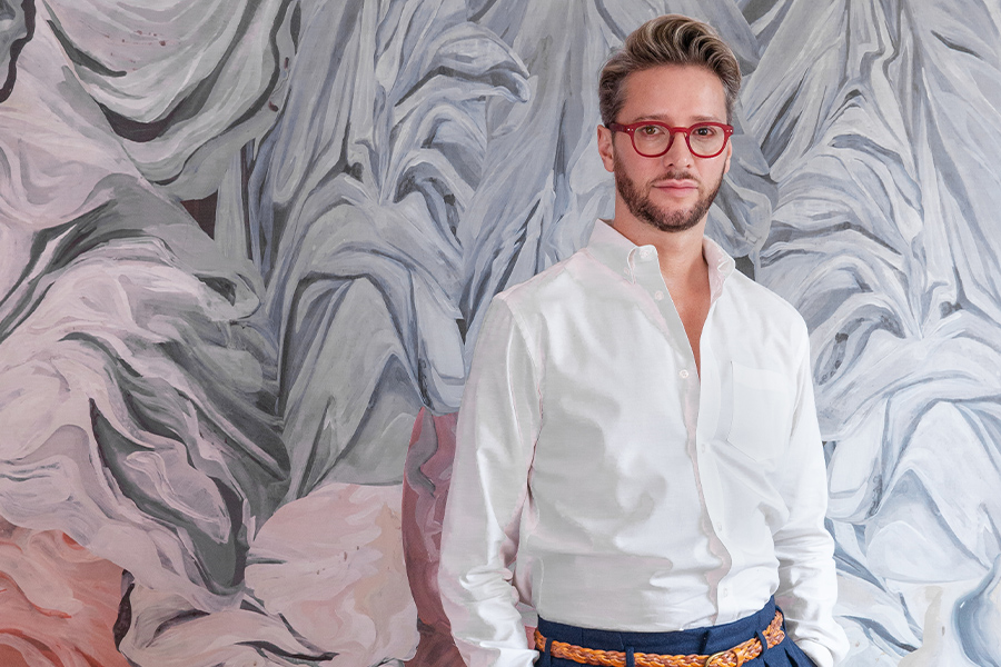 Man in a white shirt with red glasses posing against a vibrant, abstract wallpaper backdrop, exuding modern sophistication.